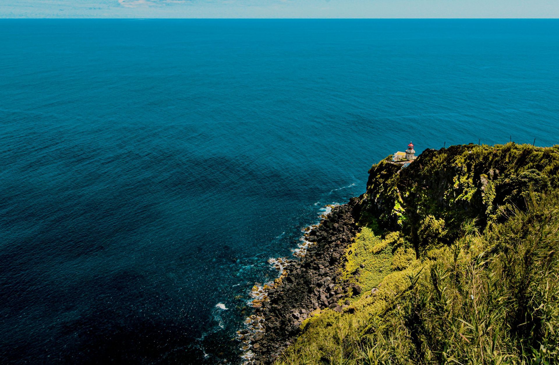 Farol em um penhasco com vista para o oceano. Água azul-escura, vegetação verde.