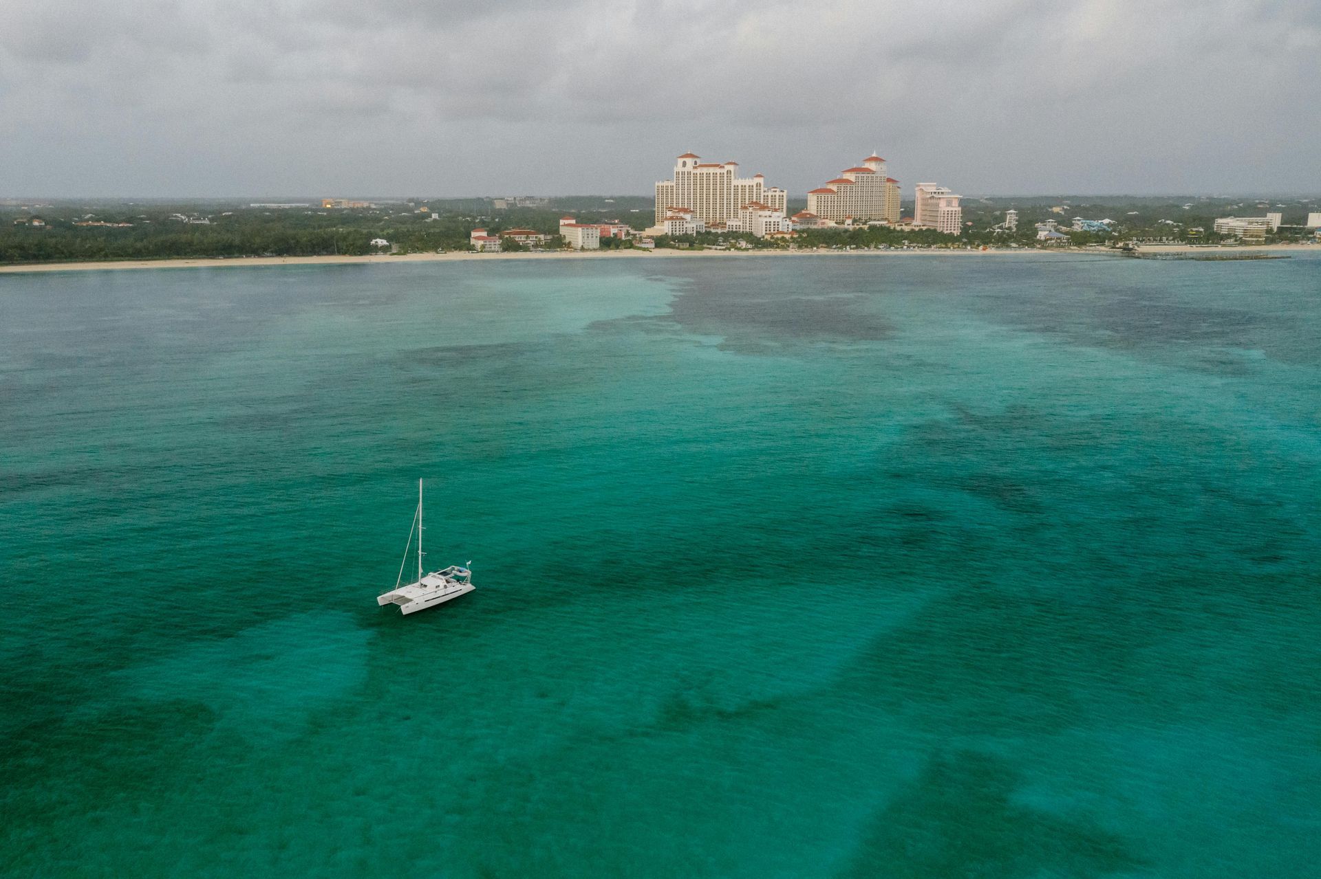 Catamaran sails on turquoise water near a coastal city under cloudy skies.