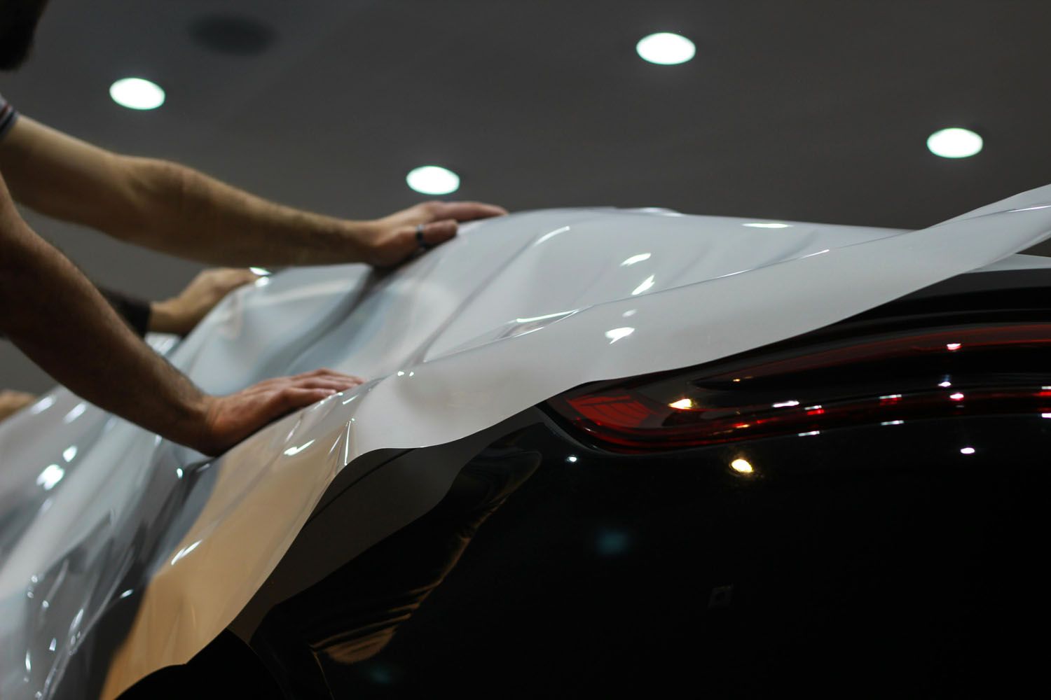 Hands carefully applying a white vinyl wrap to the curved body of a dark-colored car in a brightly lit garage.