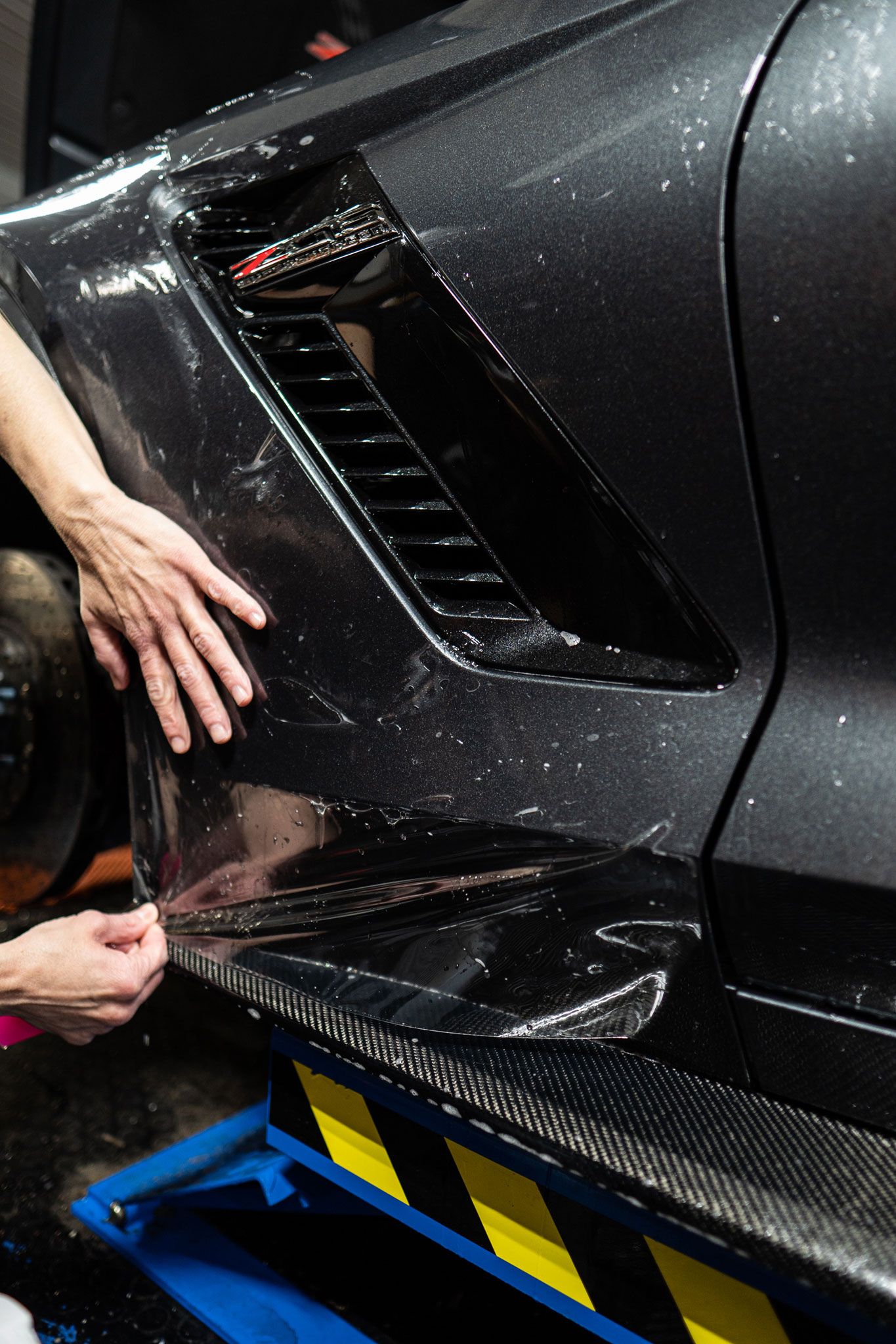 A person applies a clear paint protection film to the side panel of a dark car in a workshop.