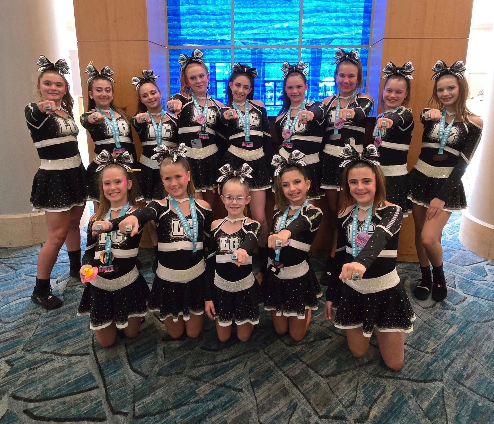Cheerleading team in black and white uniforms, posing with medals. They are standing indoors.