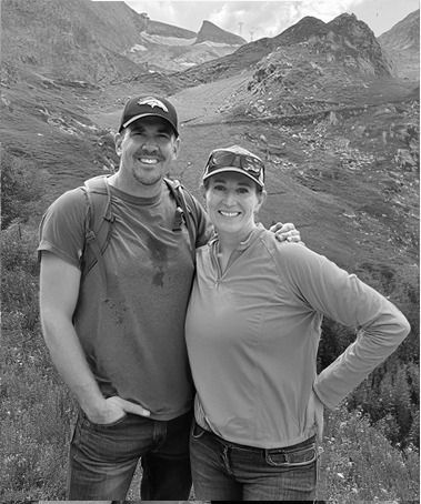 Two smiling hikers posing on a mountain trail with rocky peaks in the background