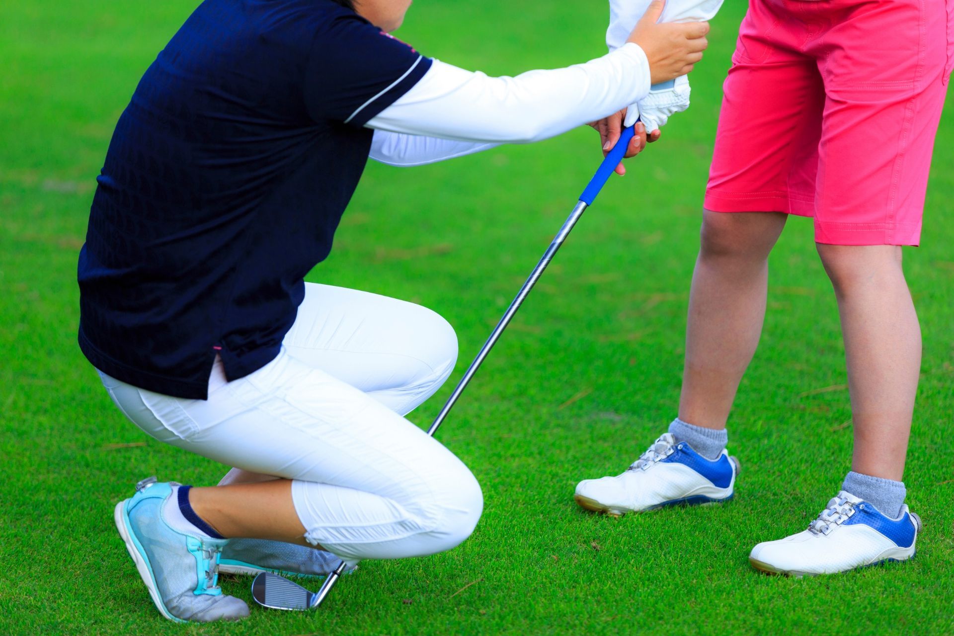 Person kneeling, adjusting the grip of a golf club for another on green grass.