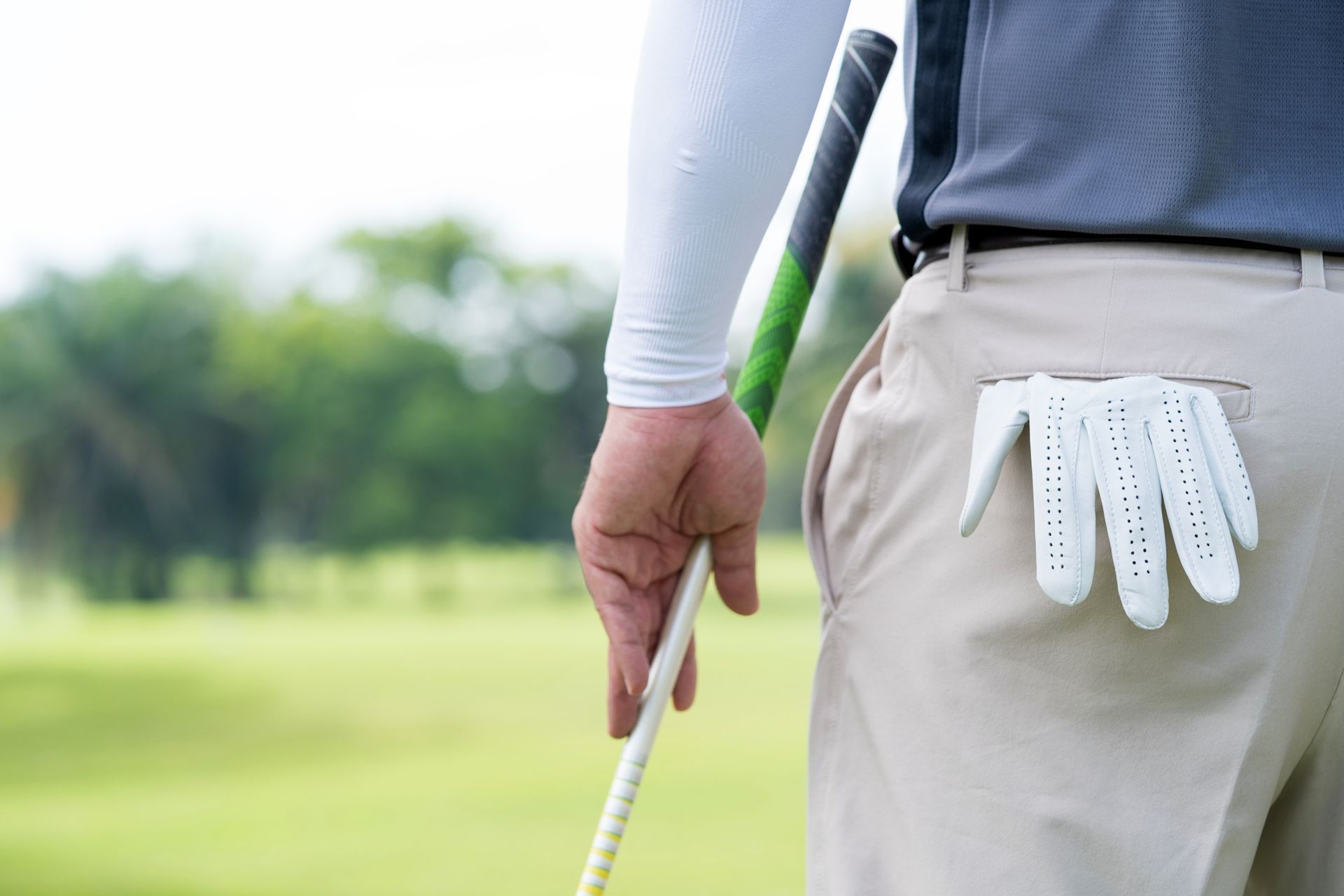 Golfer holding club on green; white glove in pocket, wearing light-colored pants and a gray shirt.