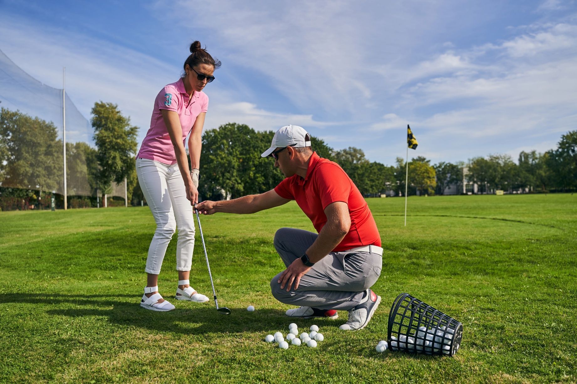Woman taking golf lesson; instructor kneels, pointing at club on green. Sunny day.