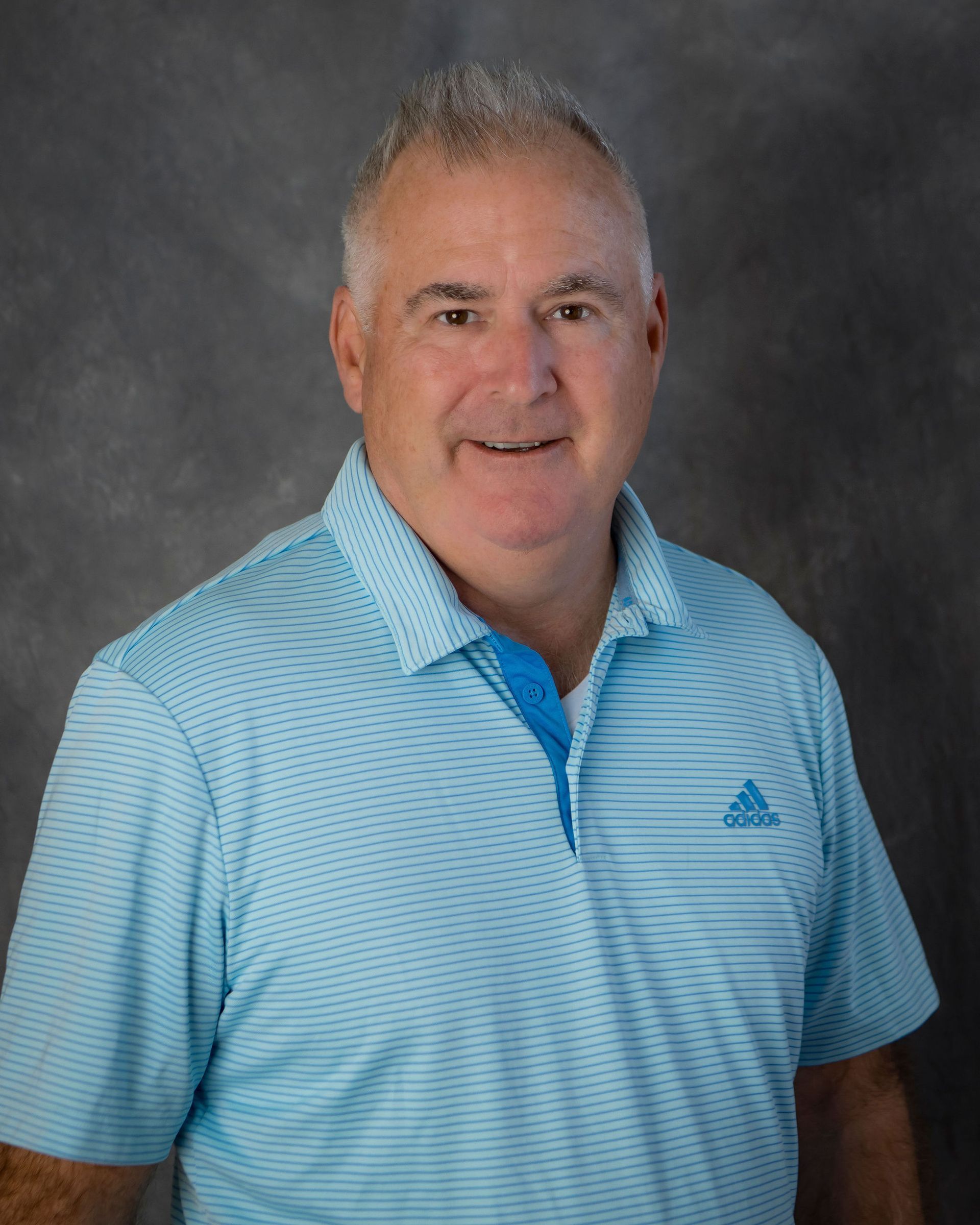A smiling person wearing a light blue, short-sleeved collared shirt with thin horizontal stripes, against a gray background.
