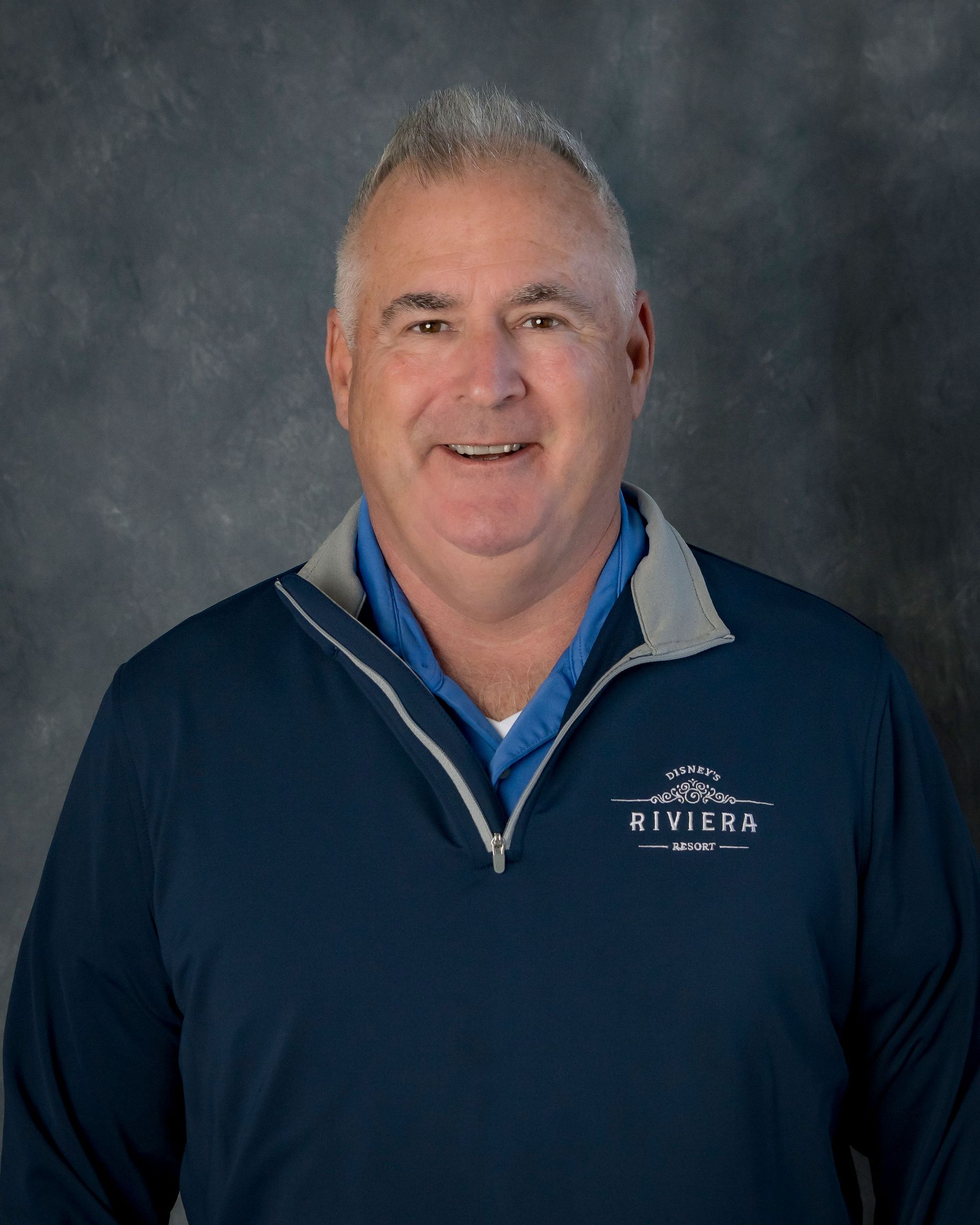 A smiling person in a blue polo shirt and a navy quarter-zip pullover with a 