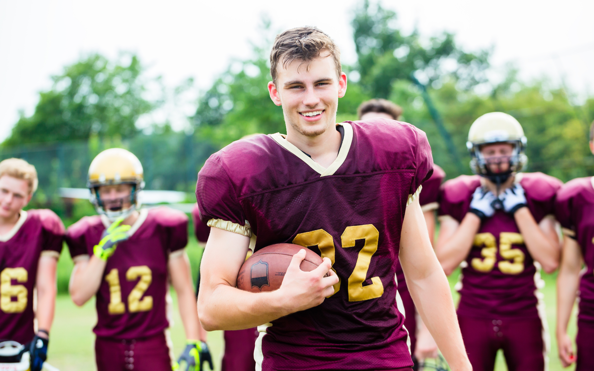 Football player in maroon jersey holding ball, smiling, surrounded by teammates on a field.