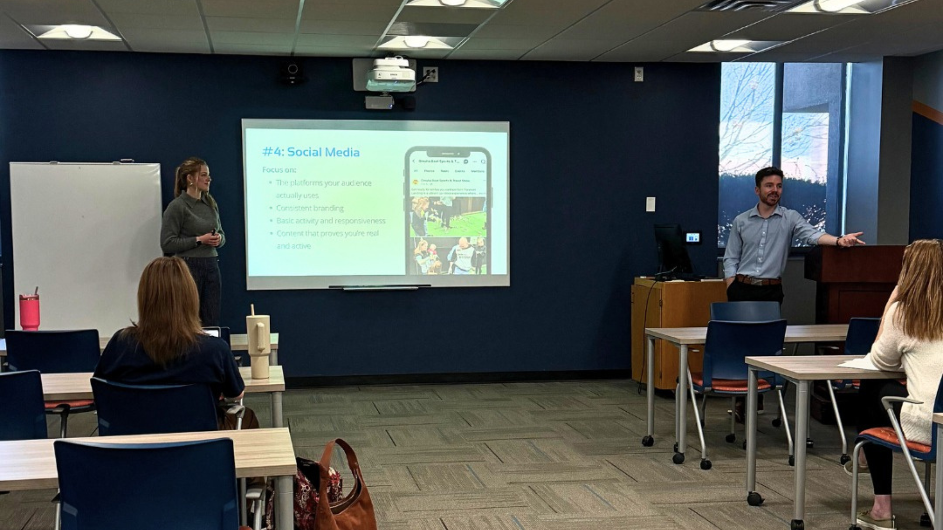 A presentation in a classroom with two speakers in front of a projection screen and several attendees seated at tables.
