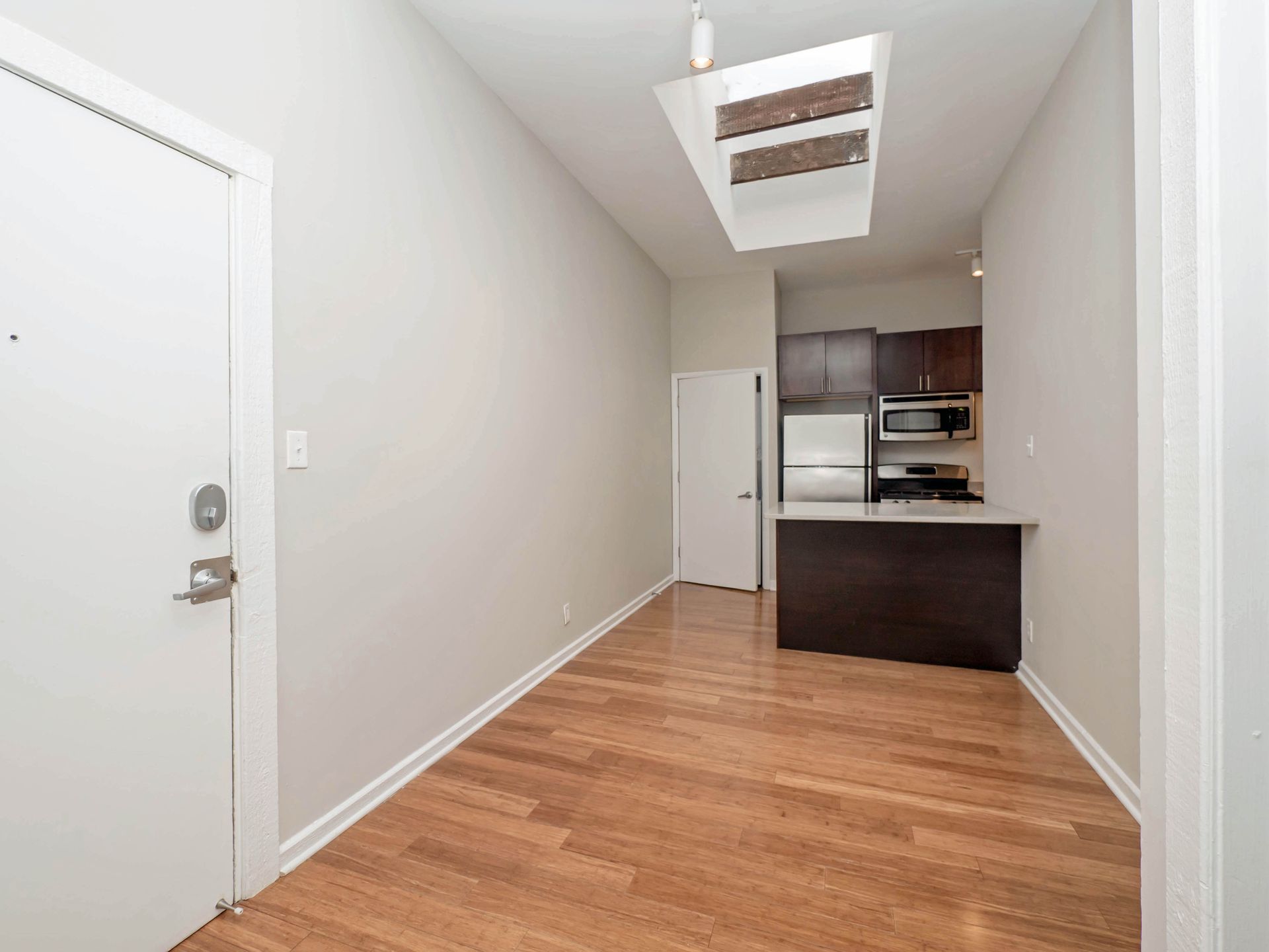 An empty apartment room with hardwood floors and a kitchen in the background.