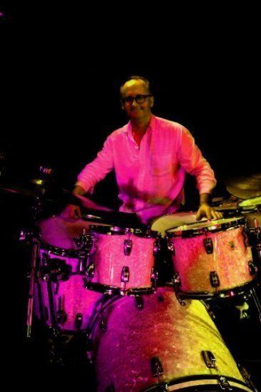 A man in a pink shirt is playing drums in a dark room.
