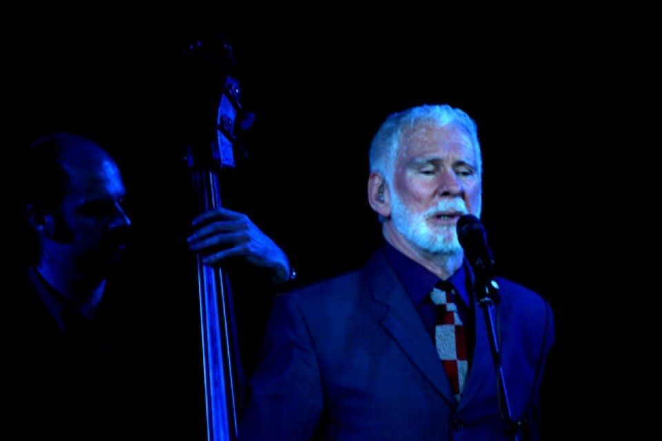 A man with a beard is singing into a microphone while another man plays a double bass.