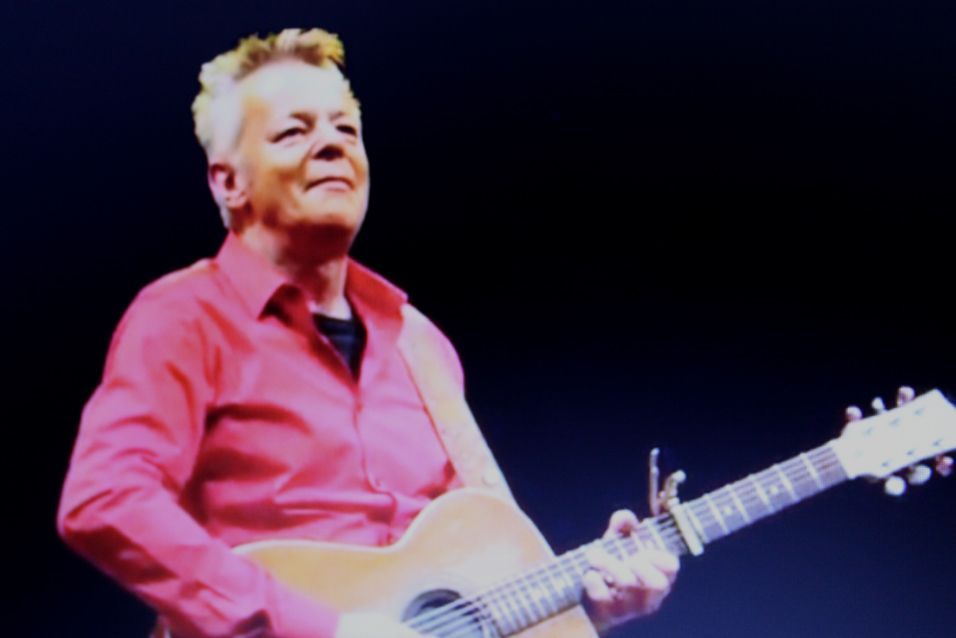 A man in a red shirt is playing an acoustic guitar
