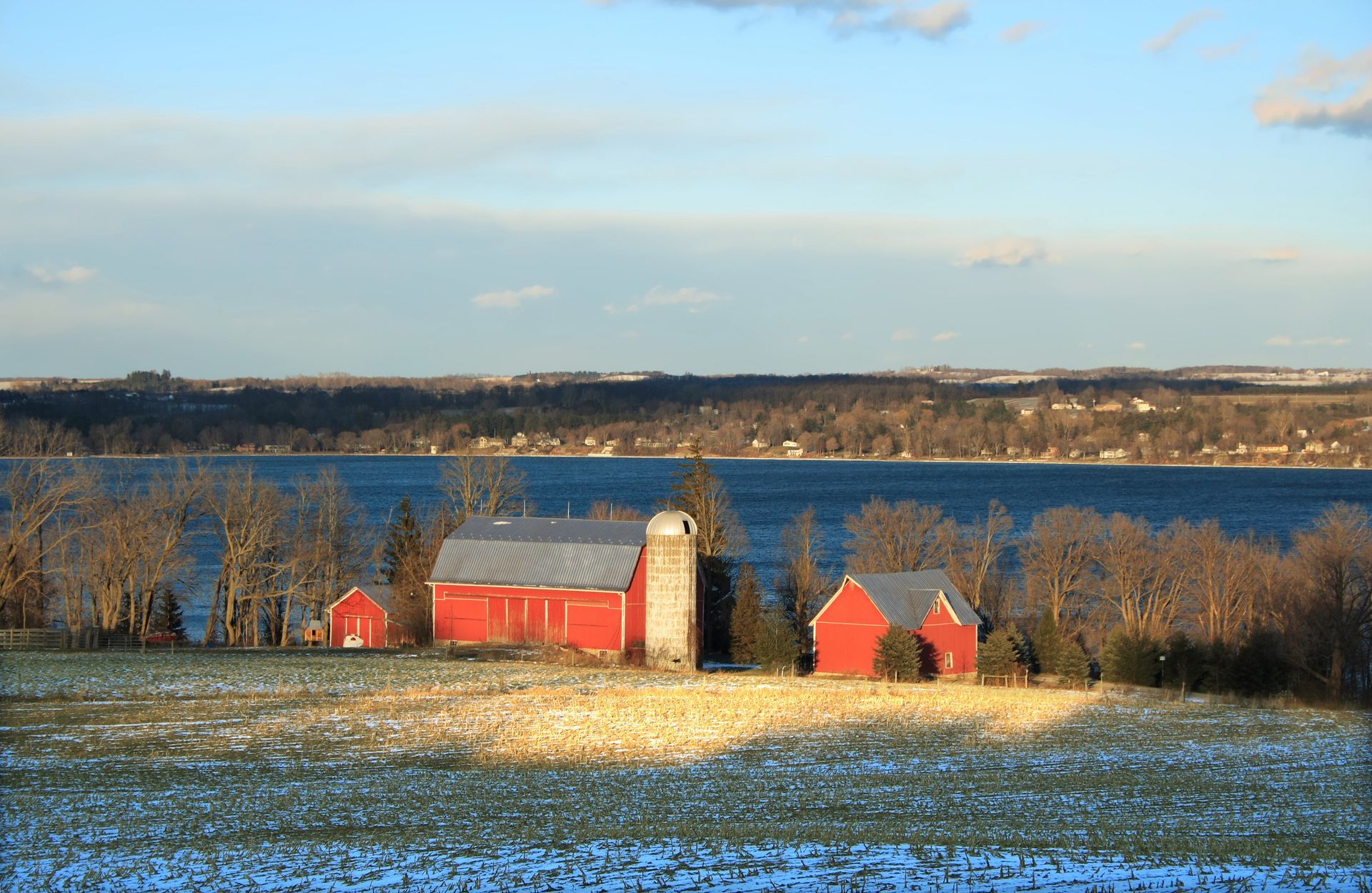 A red barn sits in the middle of a snowy field next to a body of water.