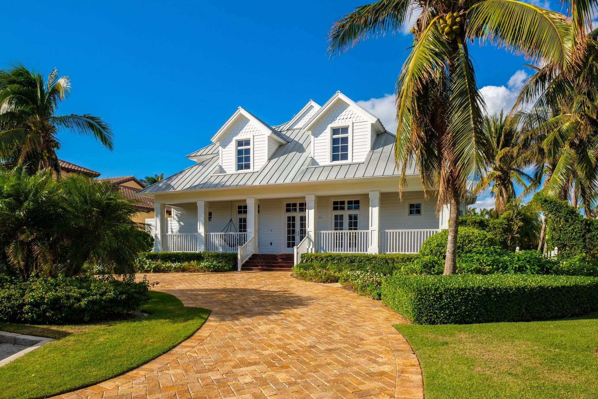 White house with a brick driveway, surrounded by green grass and palm trees under a blue sky.