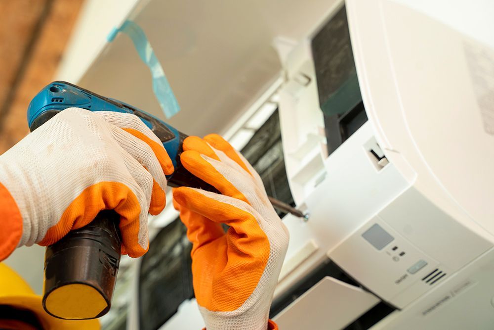 Person in orange and white gloves using a blue power drill to install an air conditioning unit.