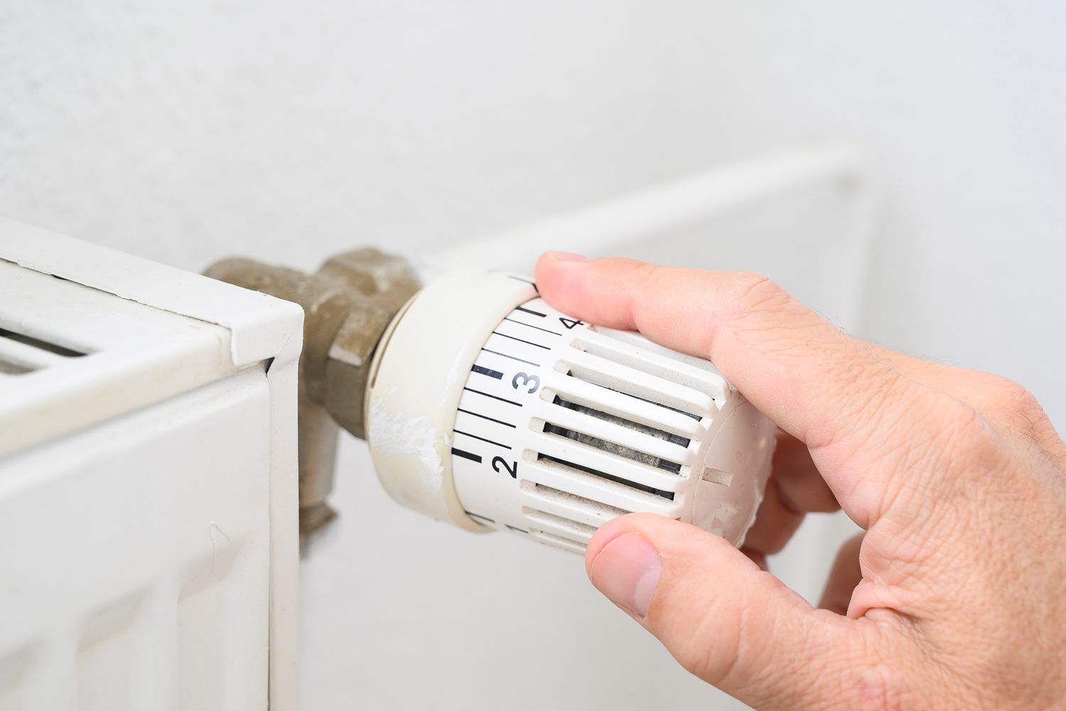 Hand turning a white thermostat dial on a radiator in a light-colored room.