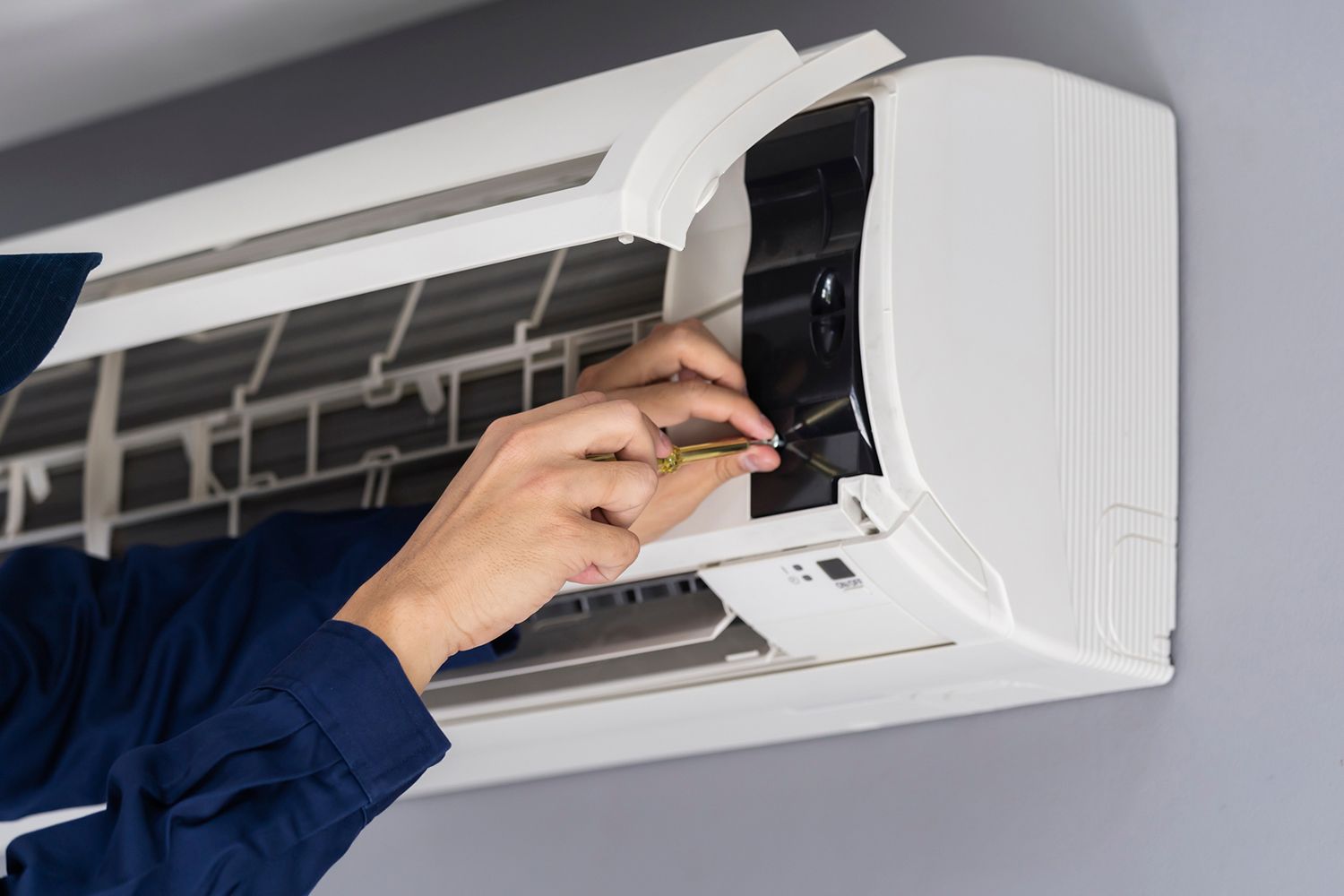 A person in blue overalls repairs a white air conditioning unit on a grey wall.