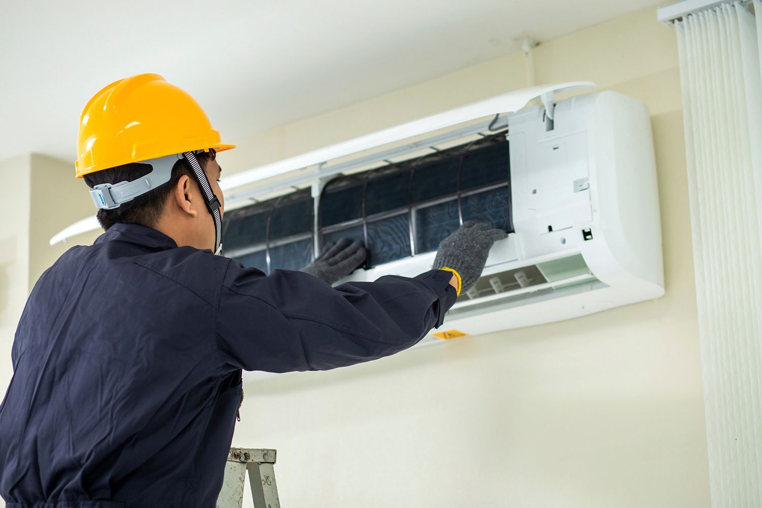 Technician in blue uniform and yellow hard hat cleaning an air conditioner filter.