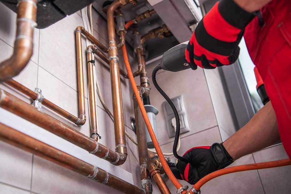 Plumber in red overalls working on copper pipes in a boiler room, using a tool, wearing gloves.