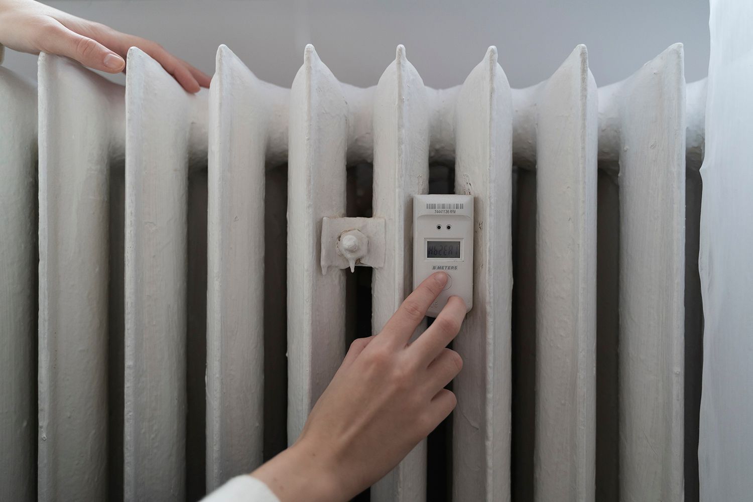 Hands adjusting a thermostat attached to a white, old-style radiator in a room.