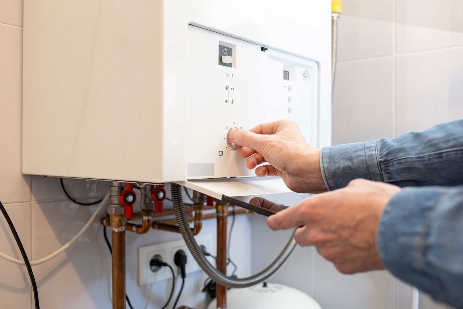 Person adjusting a white boiler, using a tool. Copper pipes and electrical cords visible.