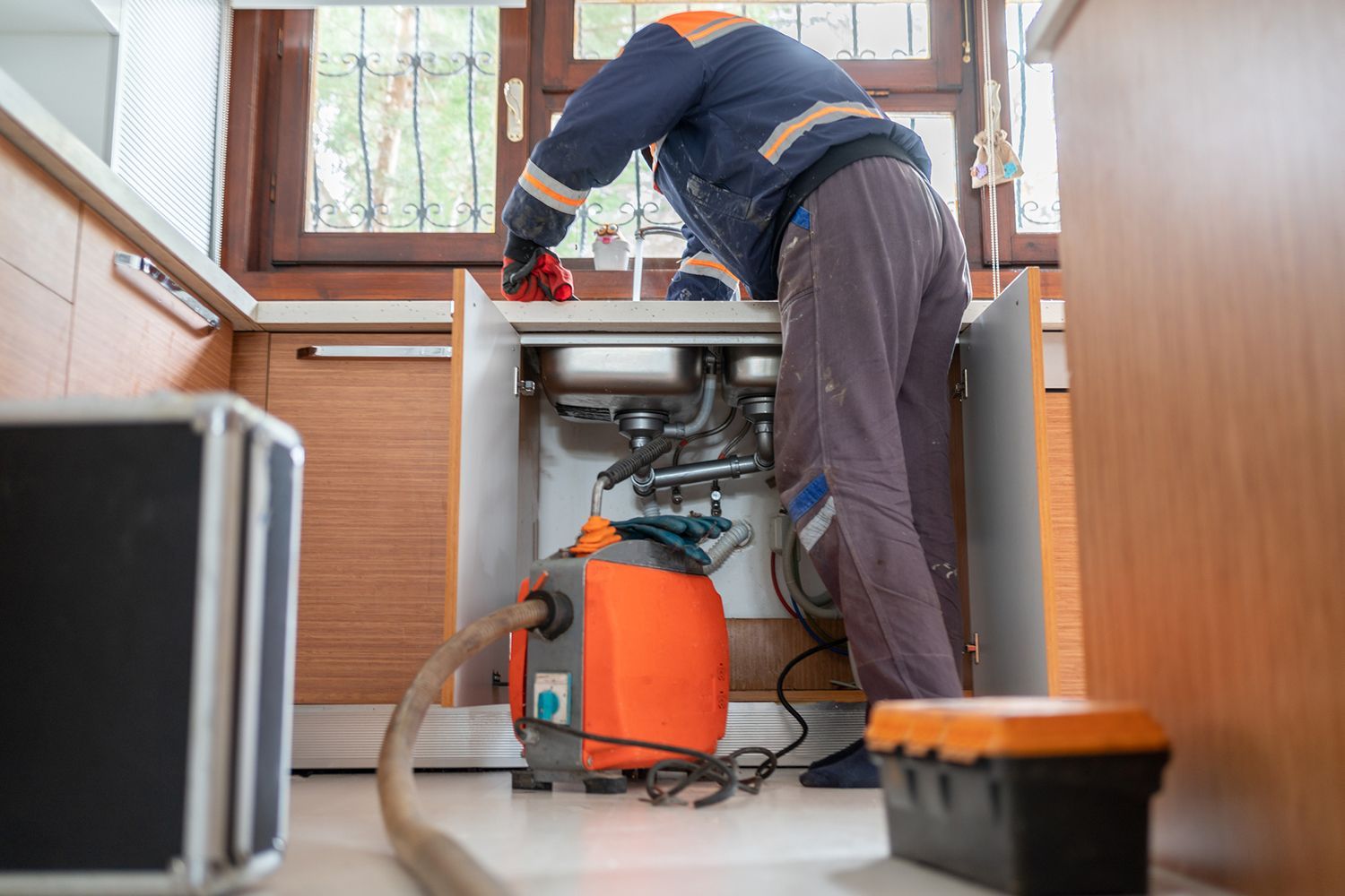 Plumber working under a kitchen sink, with a pump and toolbox visible.