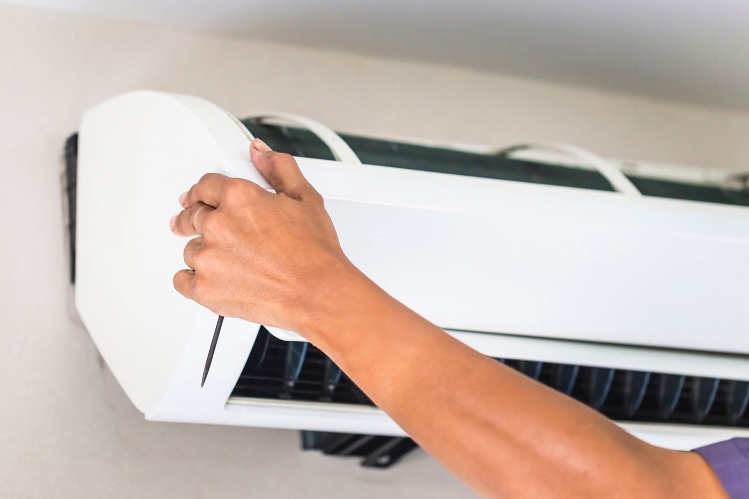 Person's hand opening the front panel of a white wall-mounted air conditioner on a white wall.