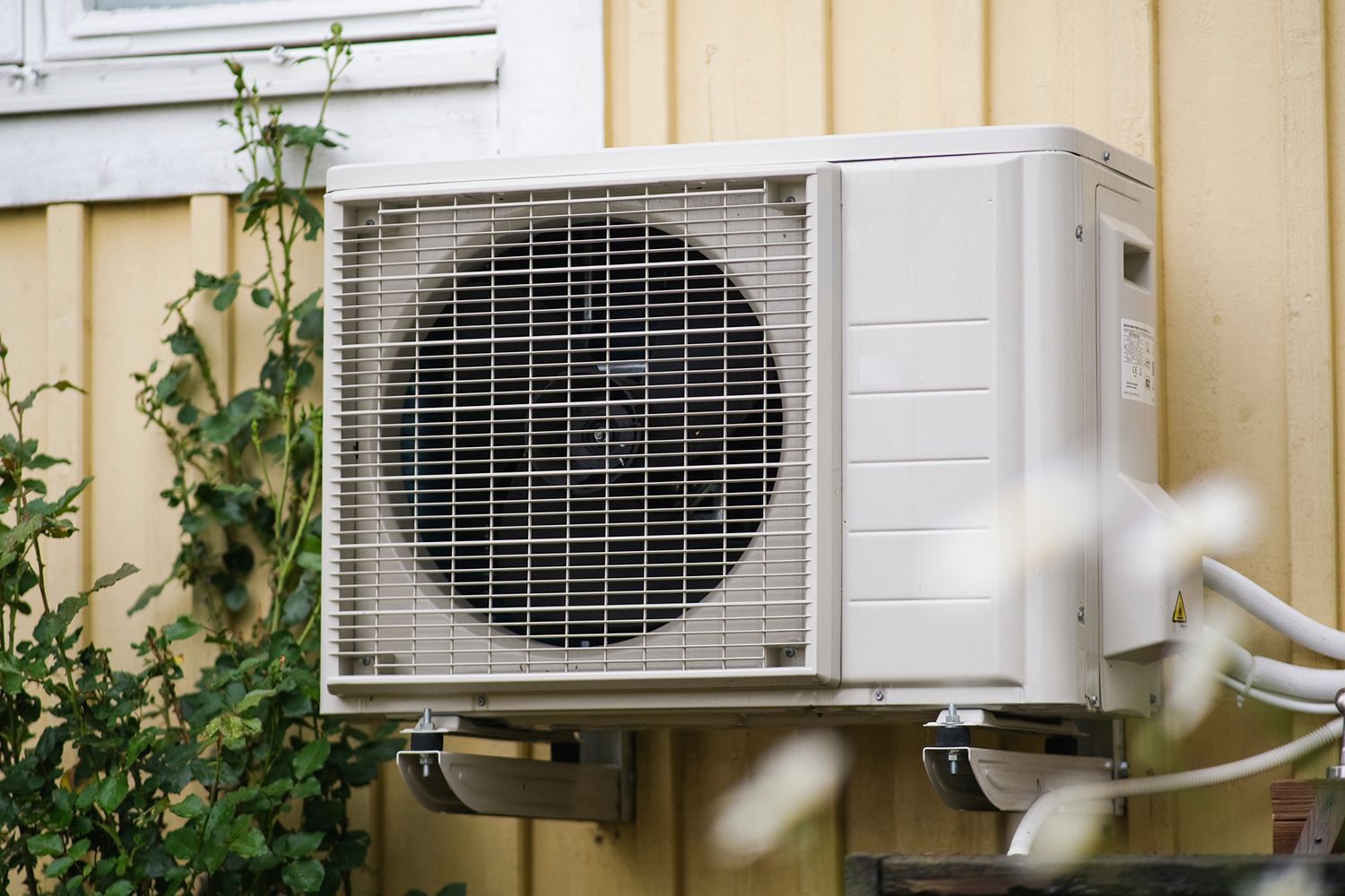 White air conditioning unit mounted on a yellow exterior wall, surrounded by greenery.