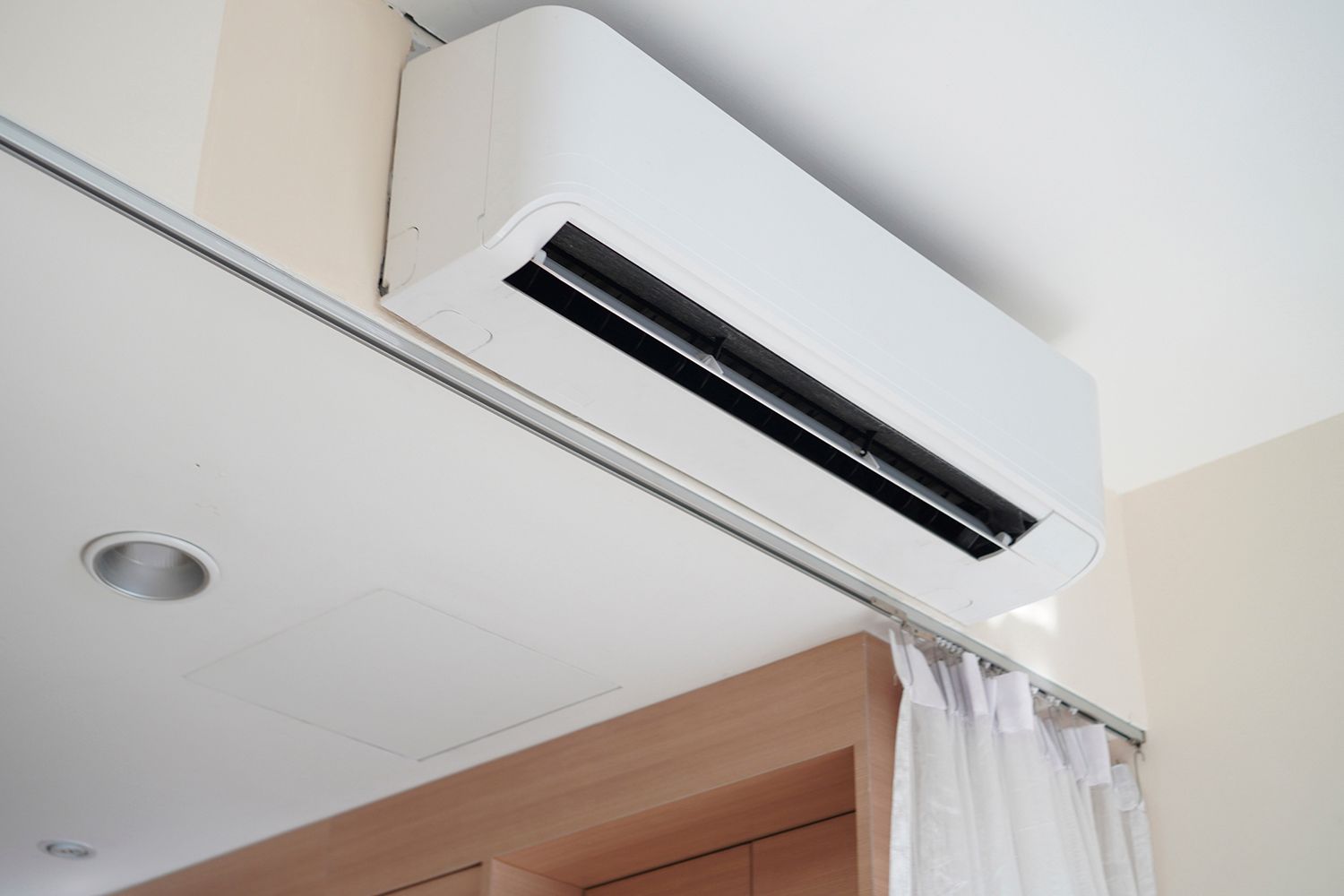 White wall-mounted air conditioner indoors, above a curtain and recessed light.