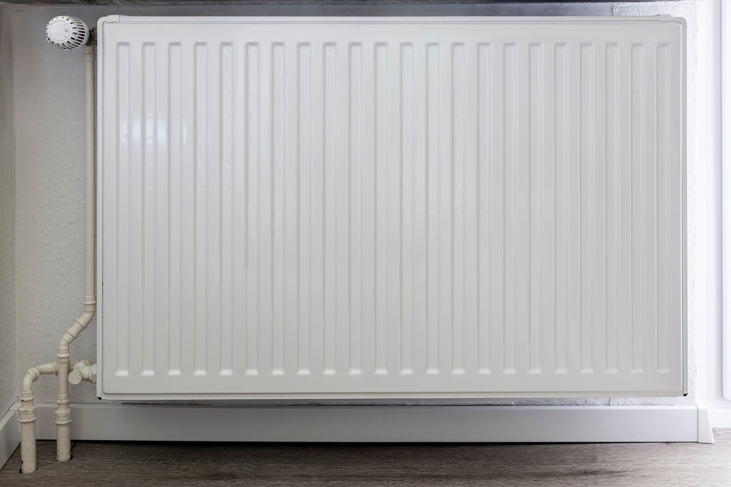 White radiator against a wall with pipes and a thermostat on the left.