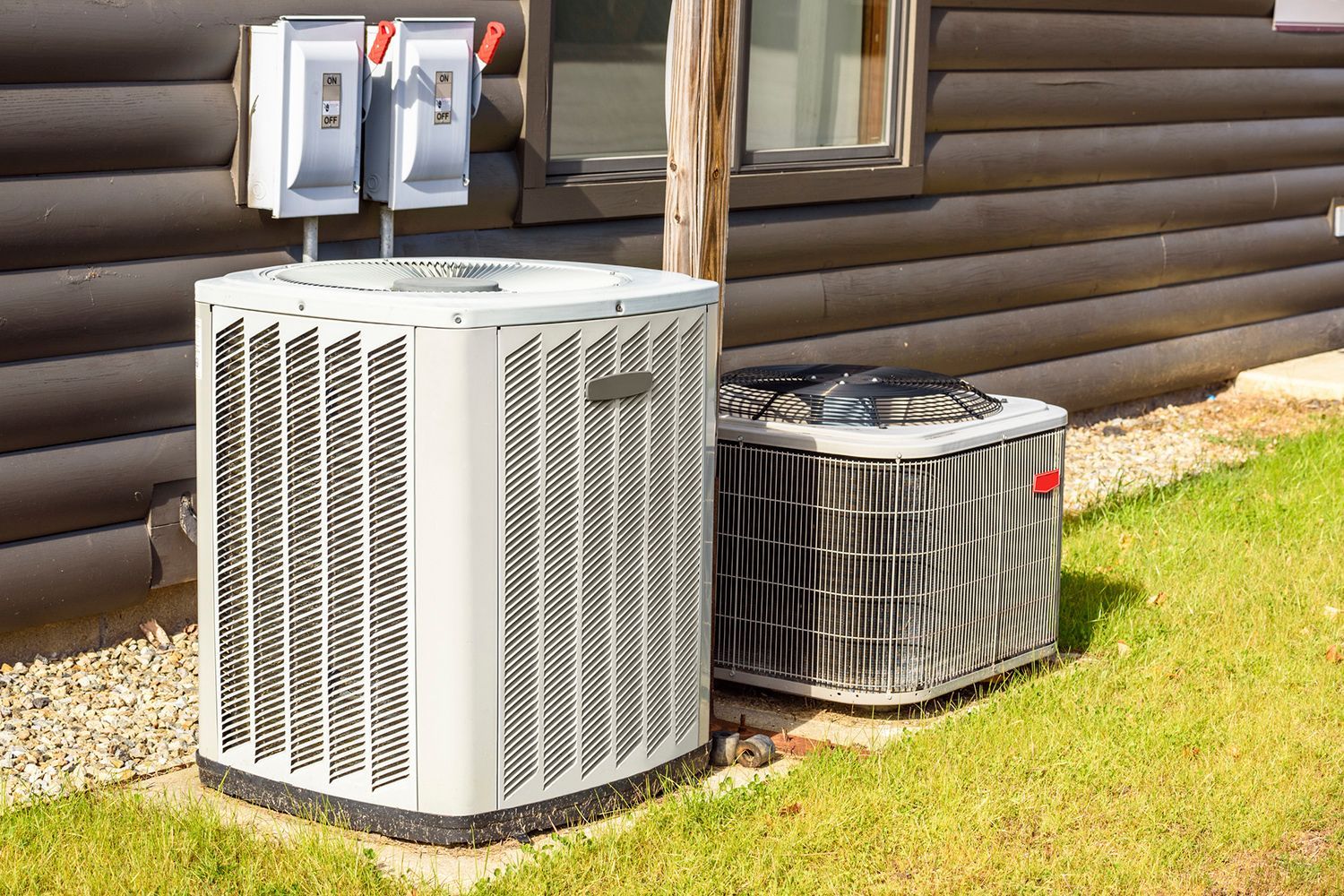 Two outdoor air conditioning units sit next to a log cabin, with electrical boxes overhead.
