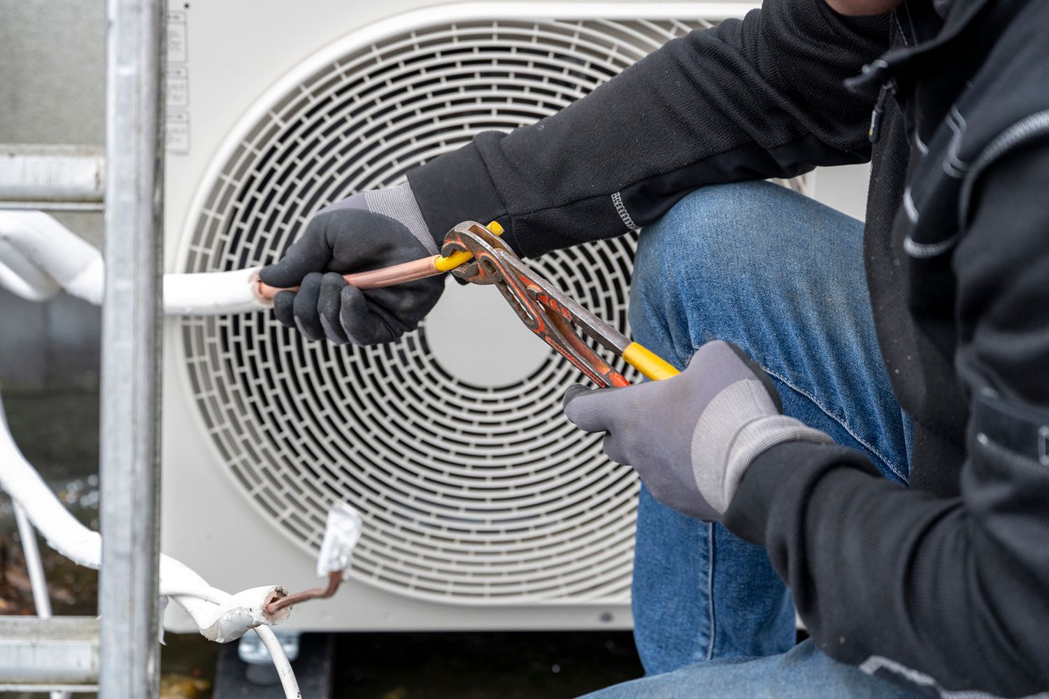 HVAC technician using pliers on copper tubing outside; wearing work gloves and jeans.