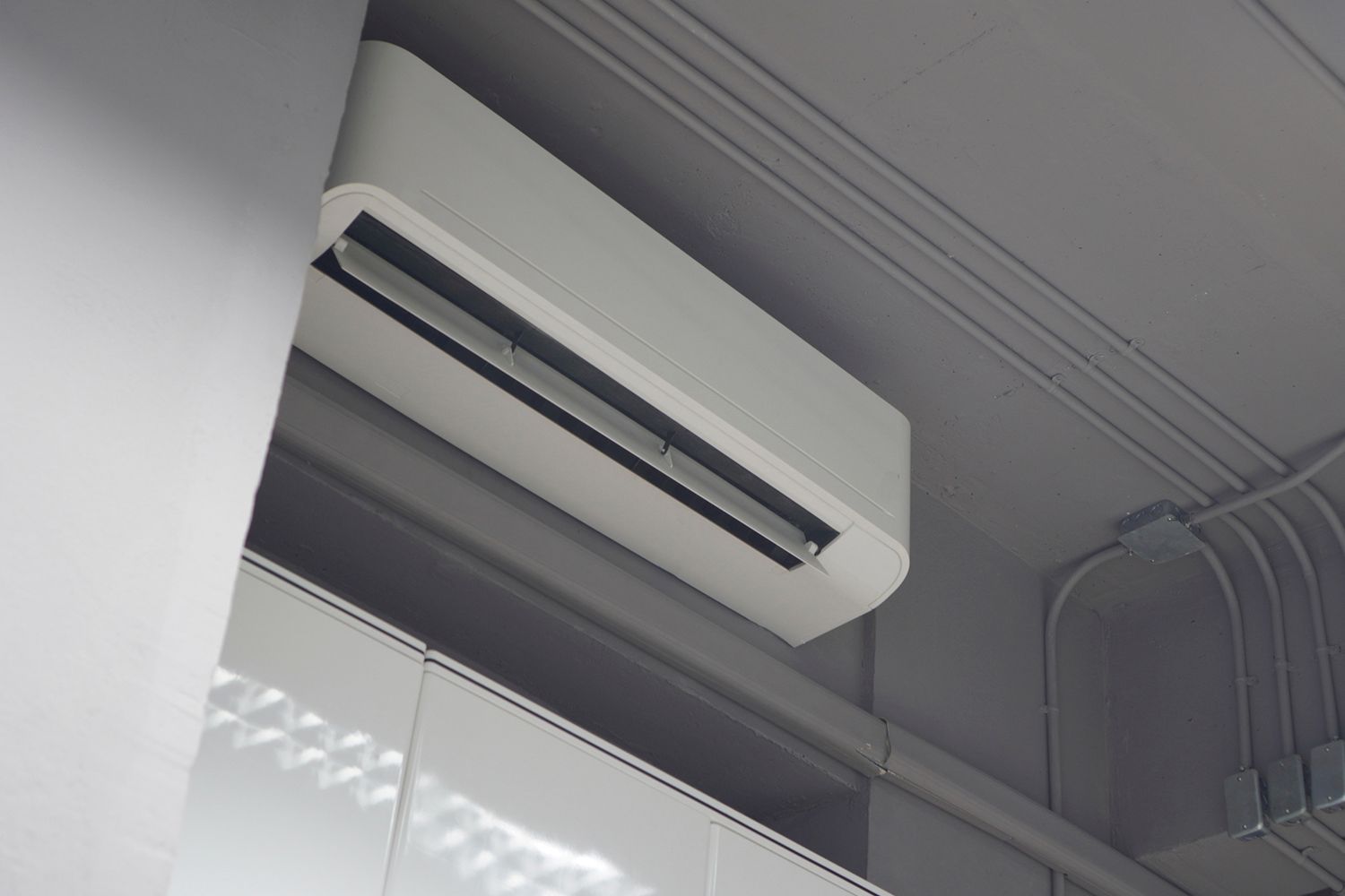 White air conditioner unit mounted on a gray wall near a ceiling with electrical conduits.