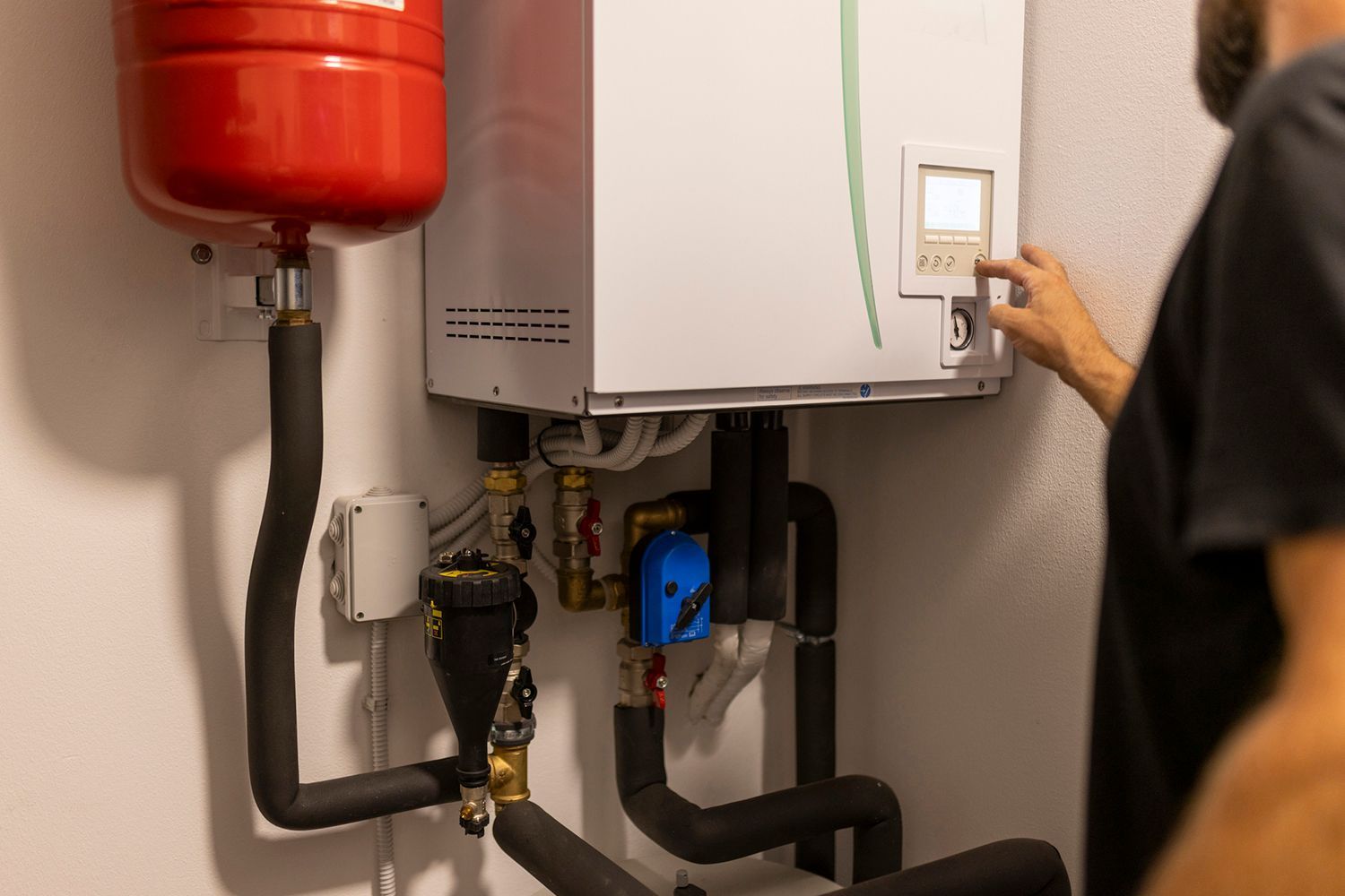 Person adjusting a white boiler on a wall, with a red tank and black pipes.