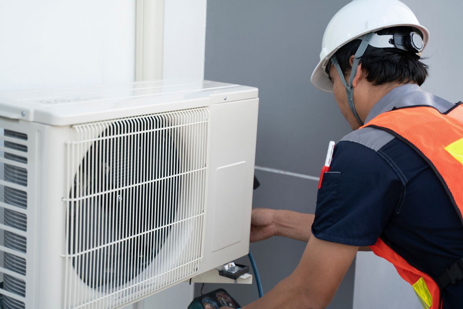 HVAC technician in hard hat and orange vest, working on an outdoor air conditioning unit.