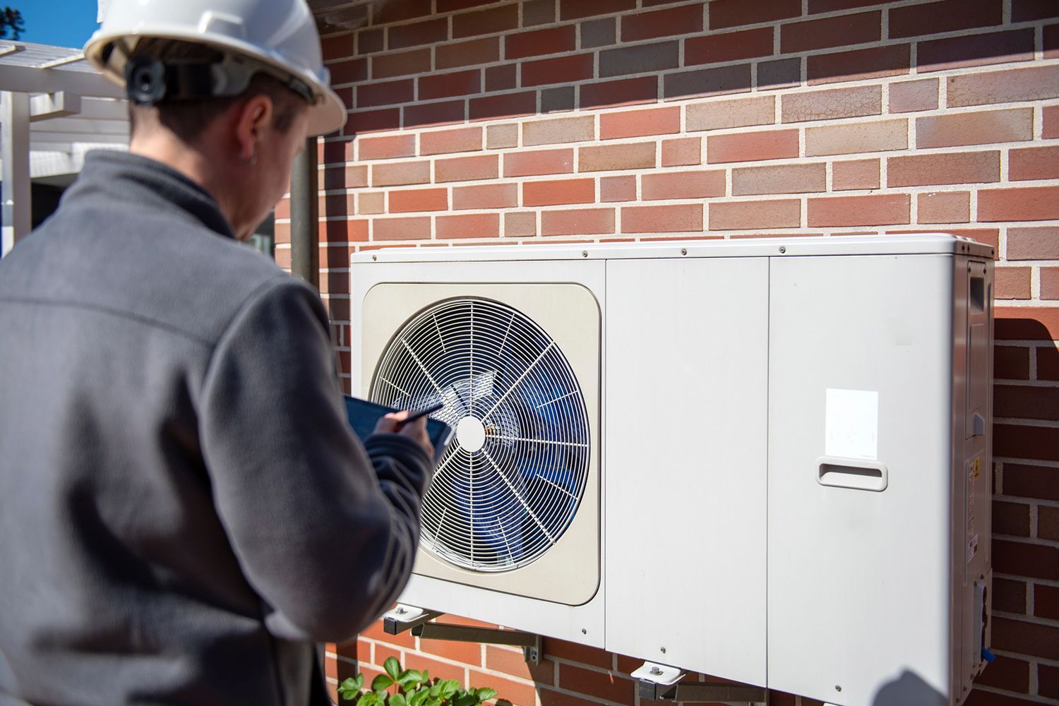 A technician in a hard hat inspecting an air conditioning unit mounted on a brick wall.