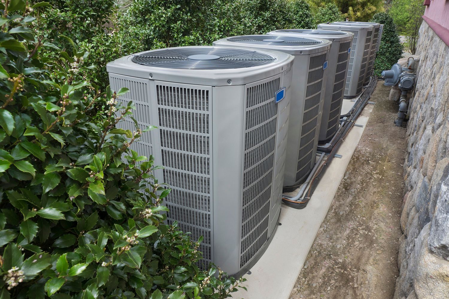 Row of air conditioning units outside a building, nestled between green bushes and a stone wall.