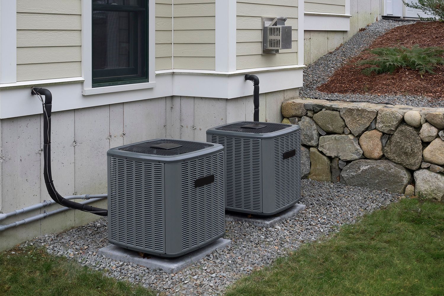 Two gray air conditioning units on a gravel bed next to a building with black pipes and a stone wall.