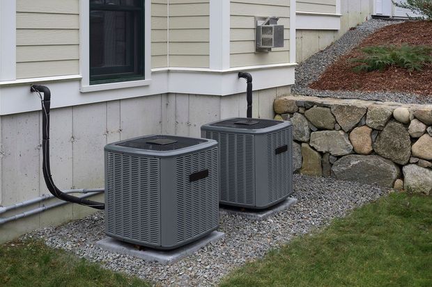 Two gray air conditioning units on a gravel bed next to a building with black pipes and a stone wall.