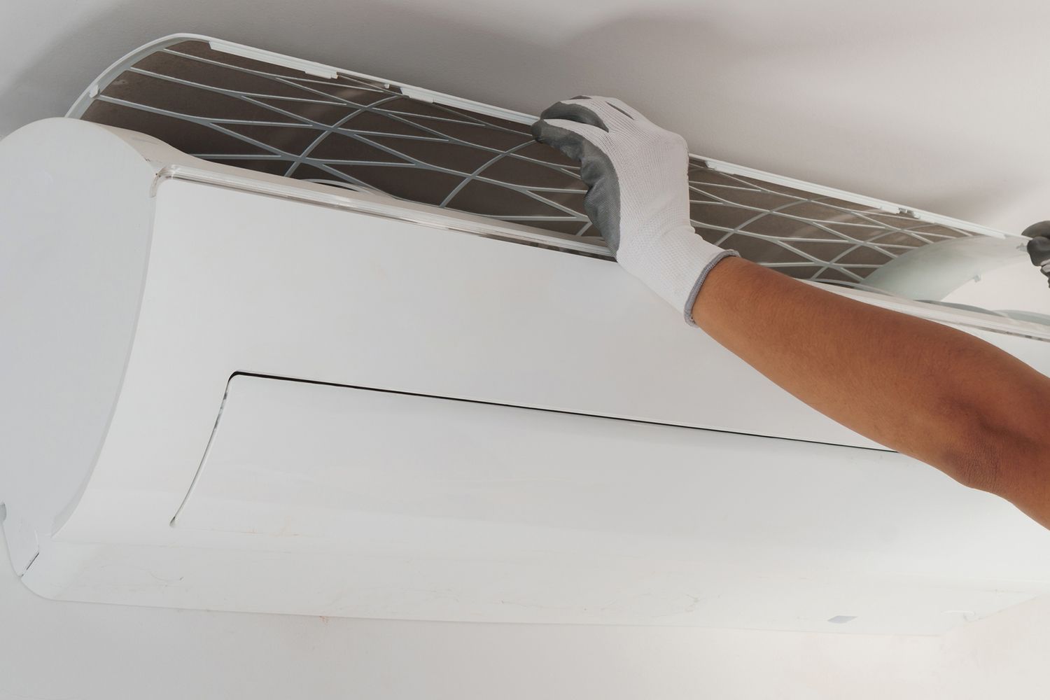 A gloved hand removes an air filter from a white air conditioner mounted on a white wall.