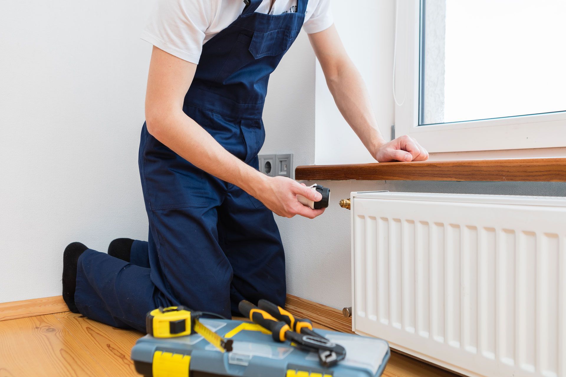A person in blue overalls kneels, repairing a white radiator near a window, tools in a toolbox.