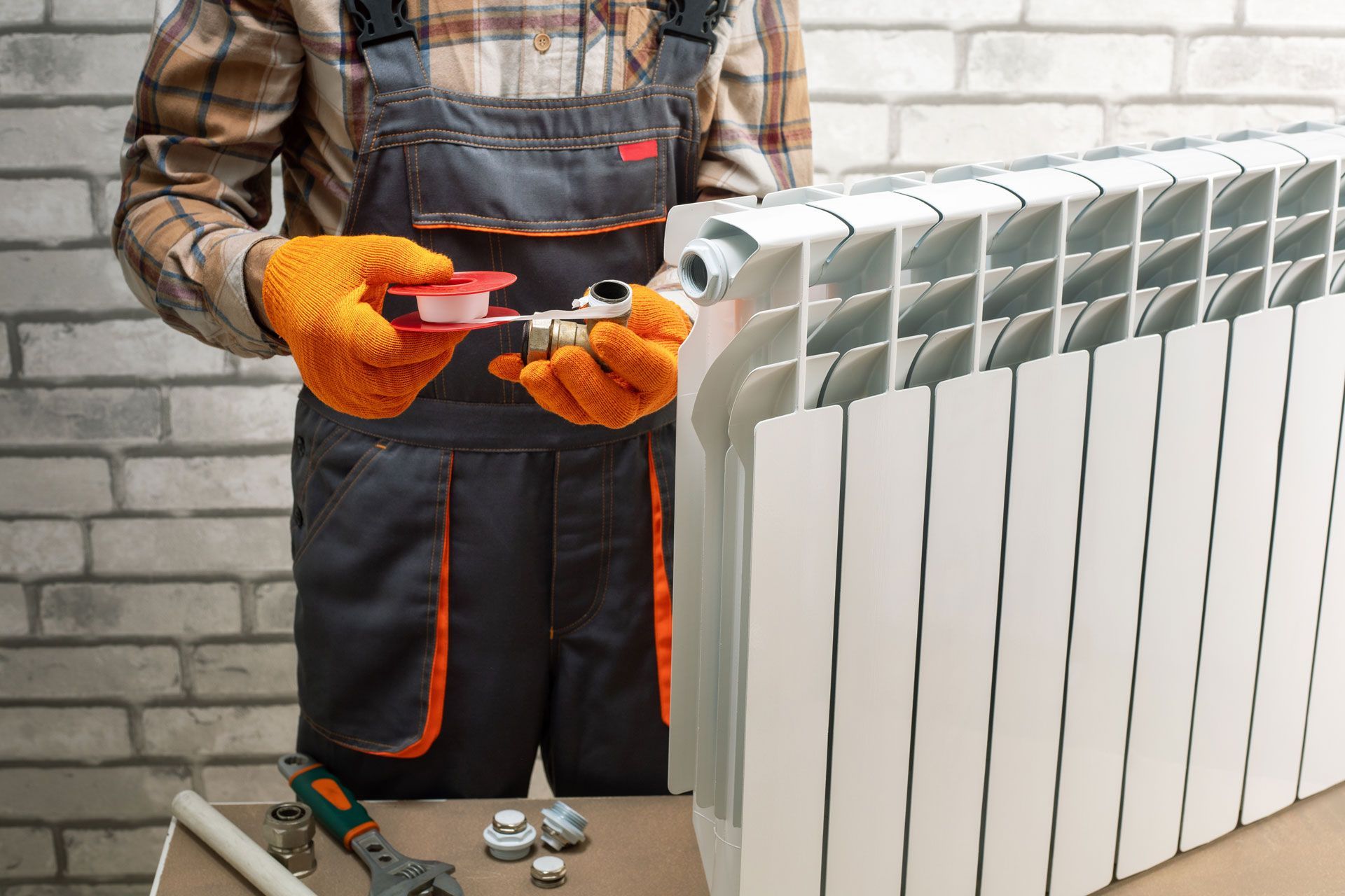 Plumber in orange gloves wraps a fitting with Teflon tape near a white radiator. Brick wall background.