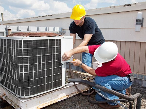 Two HVAC technicians in hard hats repair an air conditioning unit on a rooftop.