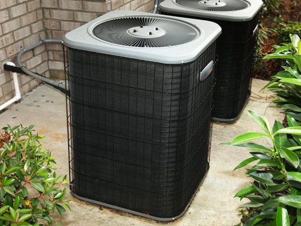 Two black air conditioning units outside on a concrete slab, surrounded by greenery.