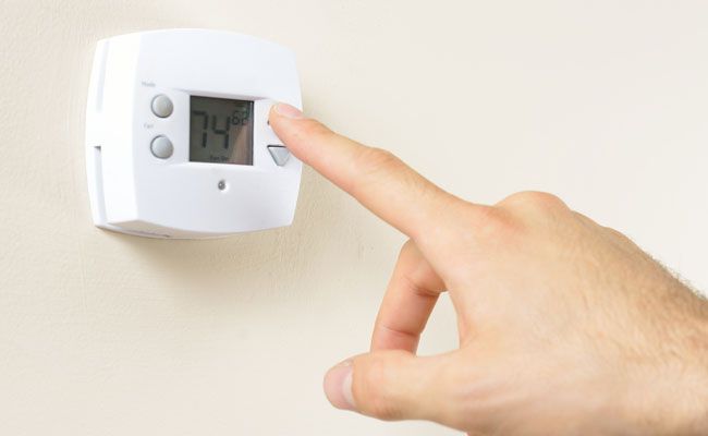 Hand adjusting a digital thermostat set to 74 degrees Fahrenheit, mounted on a beige wall.