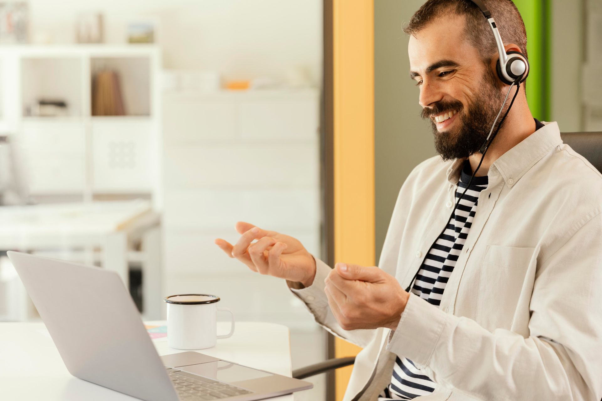 Hombre sonriendo con auriculares puestos frente a su computadora portátil, gesticulando durante una videollamada en una luminosa oficina en casa.