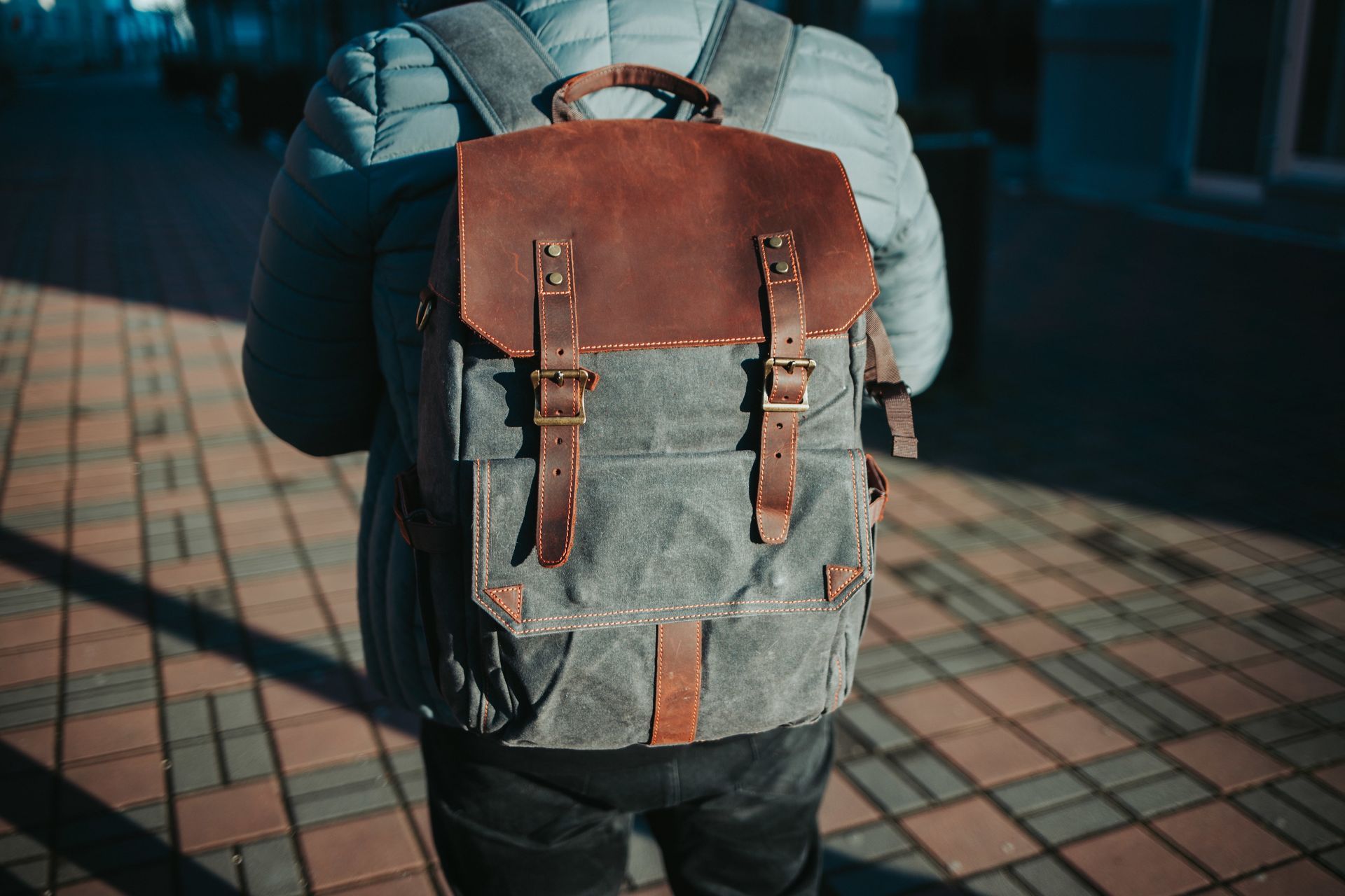 Persona con mochila gris y marrón caminando por una acera de baldosas.