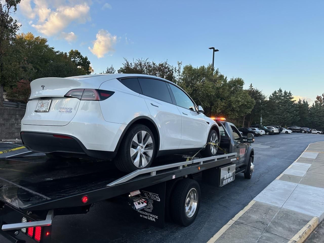 White Tesla Model Y on a tow truck, roadside with other cars and trees. Evening sky.