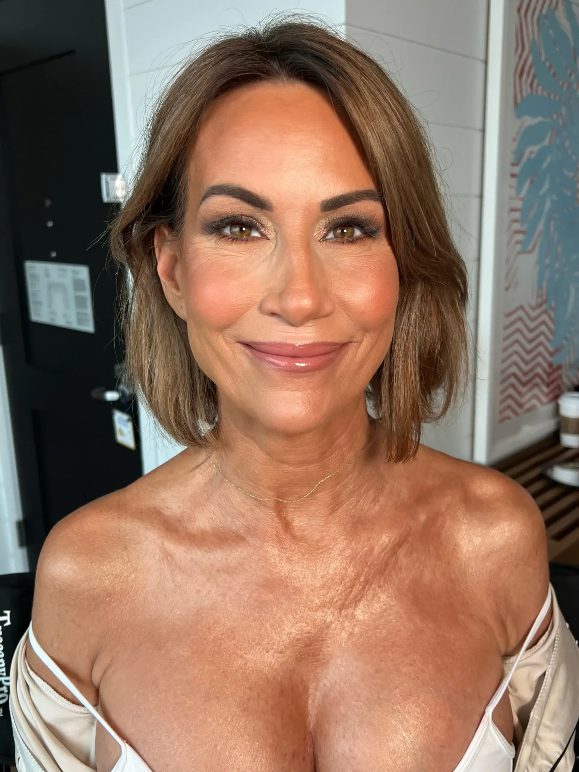 A woman with brown hair and makeup smiles. She wears a light-colored top with bare shoulders, appearing in an indoor setting.